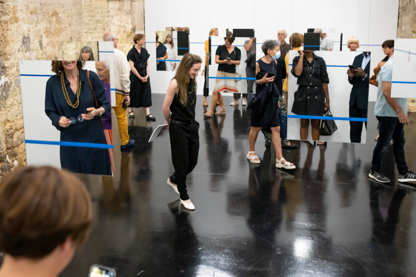 Dans une galerie, un groupe de visiteurs déambule au milieu de miroirs suspendus et barrés horizontalement par un ruban d'adhésif bleu.