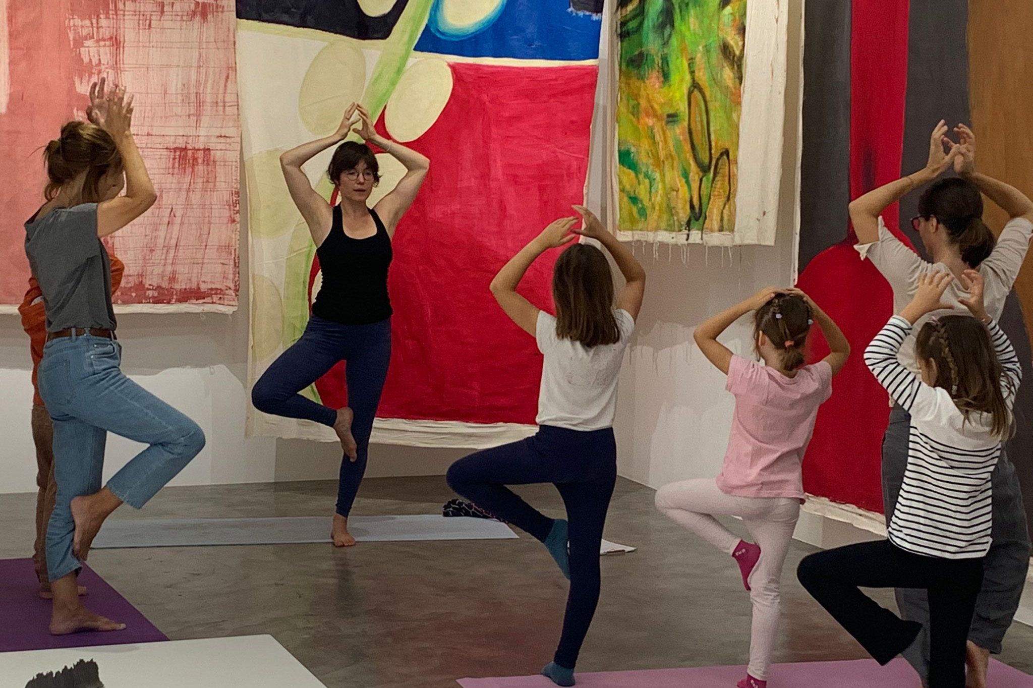 Un groupe constitué d'adultes et d'enfants font du yoga devant des peintures abtraites colorées à dominante rouge
