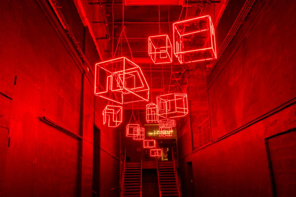 Un large couloir baigné d'un lumière rouge intense. En face, deux escaliers en métal de type industriel. Au plafond des suspensions en néon rouge forment des volumes comme pour représenter des habitations. 