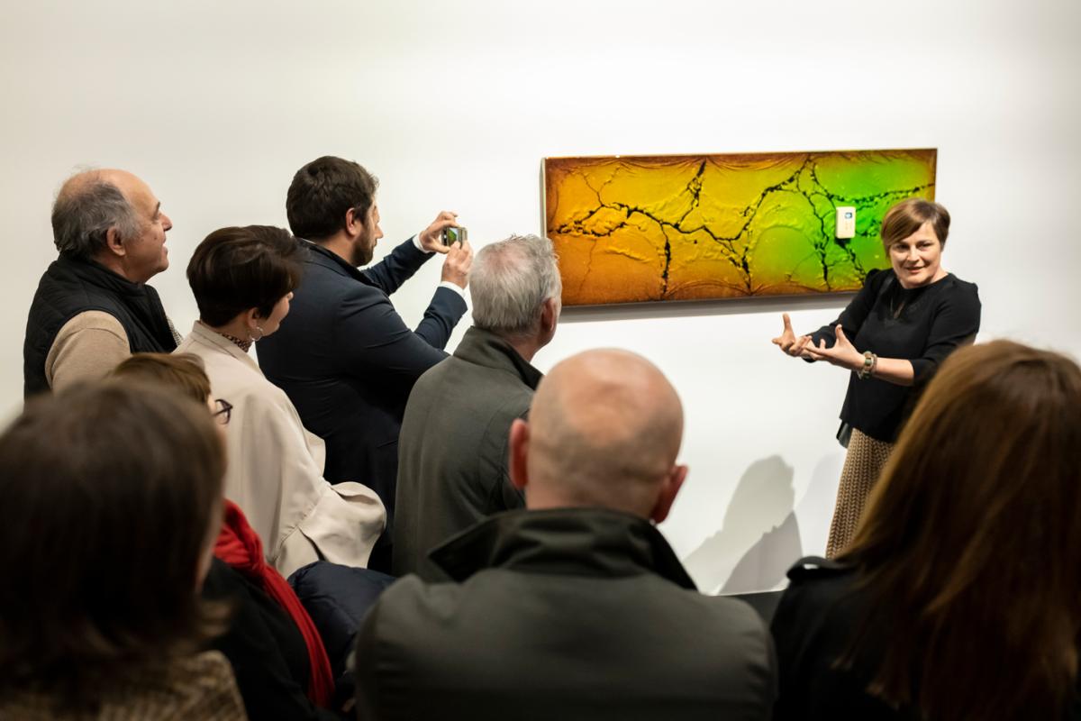 Face a un groupe de personnes, la directrice du musée présente une oeuvre aux couleur verte et jeune accorichée sur un mur blanc. Sur l'oeuvre on distingue des craquelures et on aperçoit un capteur.