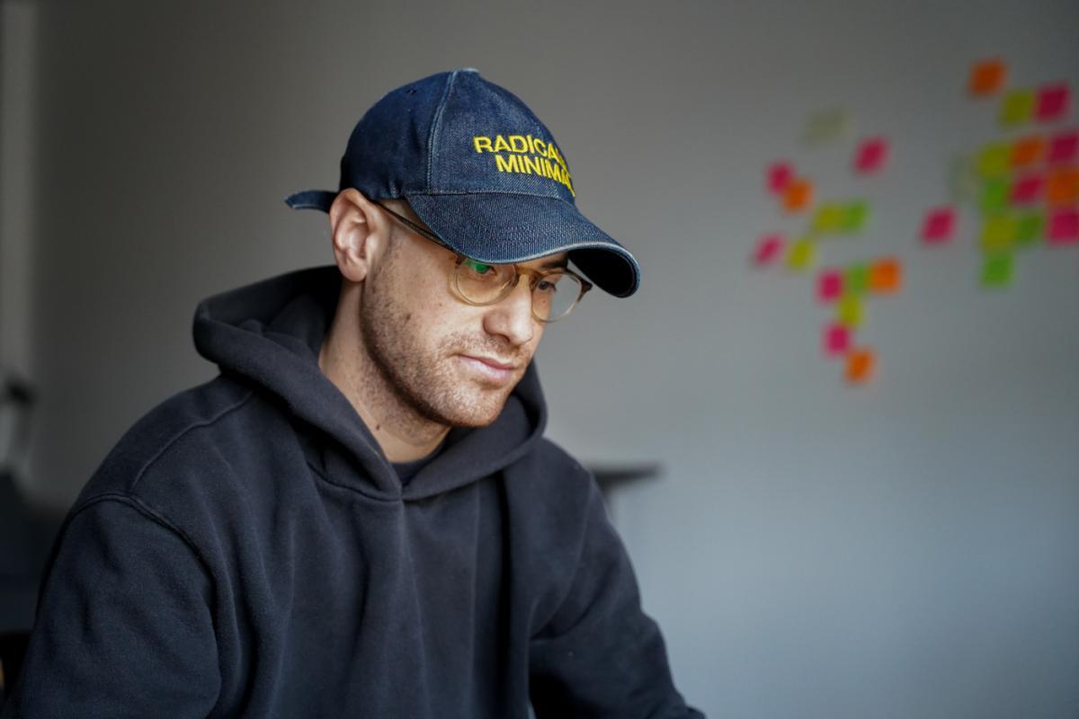 Portrait of a young man wearing a grey hooded sweatshirt and a dark blue cap. A constellation of coloured post-its can be seen in the background on a white wall. 
