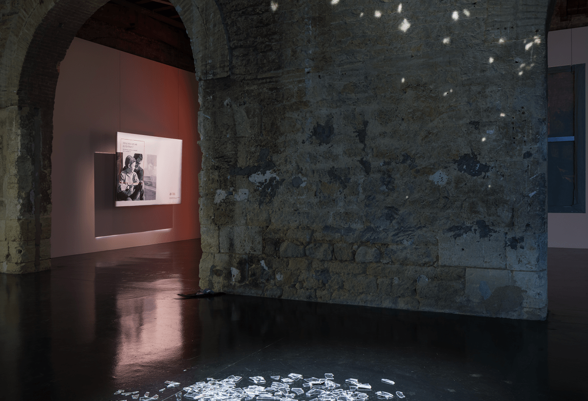 Des éclats de miroirs se reflètent sur les murs de l'exposition dans une salle sombre de l'exposition. On aperçoit une peinture de l'artiste au loin.