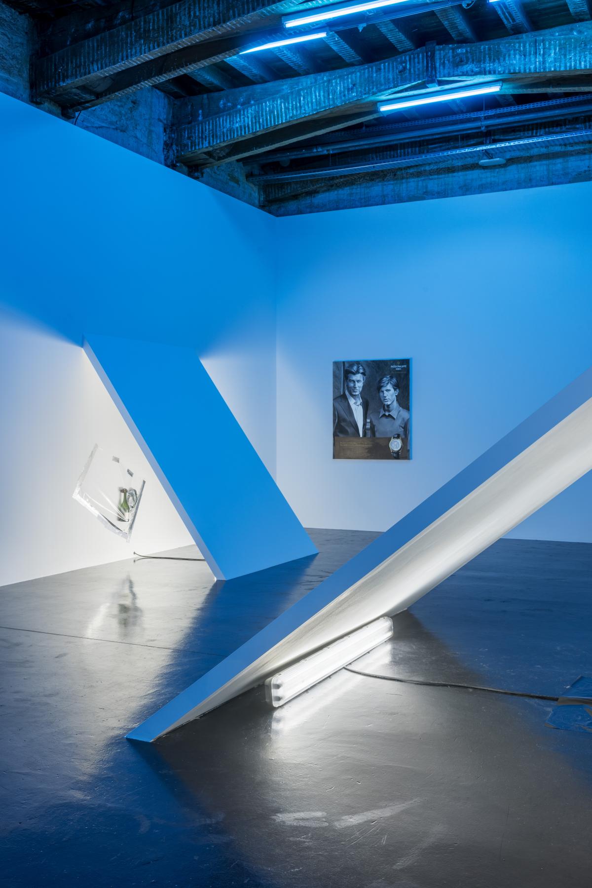 La salle de l’exposition est baignée dans une lumière qui teinte l’espace en bleu. Des extensions des murs font obstacle, empêchant ainsi une circulation libre dans l'espace. Sous ces pans de murs obliques, des bandes de tissus et déchets sont recouverts de films plastifiés. On aperçoit une toile au fond de la salle.