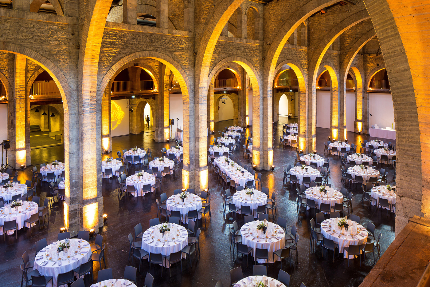 Sous de hautes voutes en pierre sublimées par un éclairage doré, un ensemble de plus d'une quarantaine de tables sont napées, dressées et fleuries.