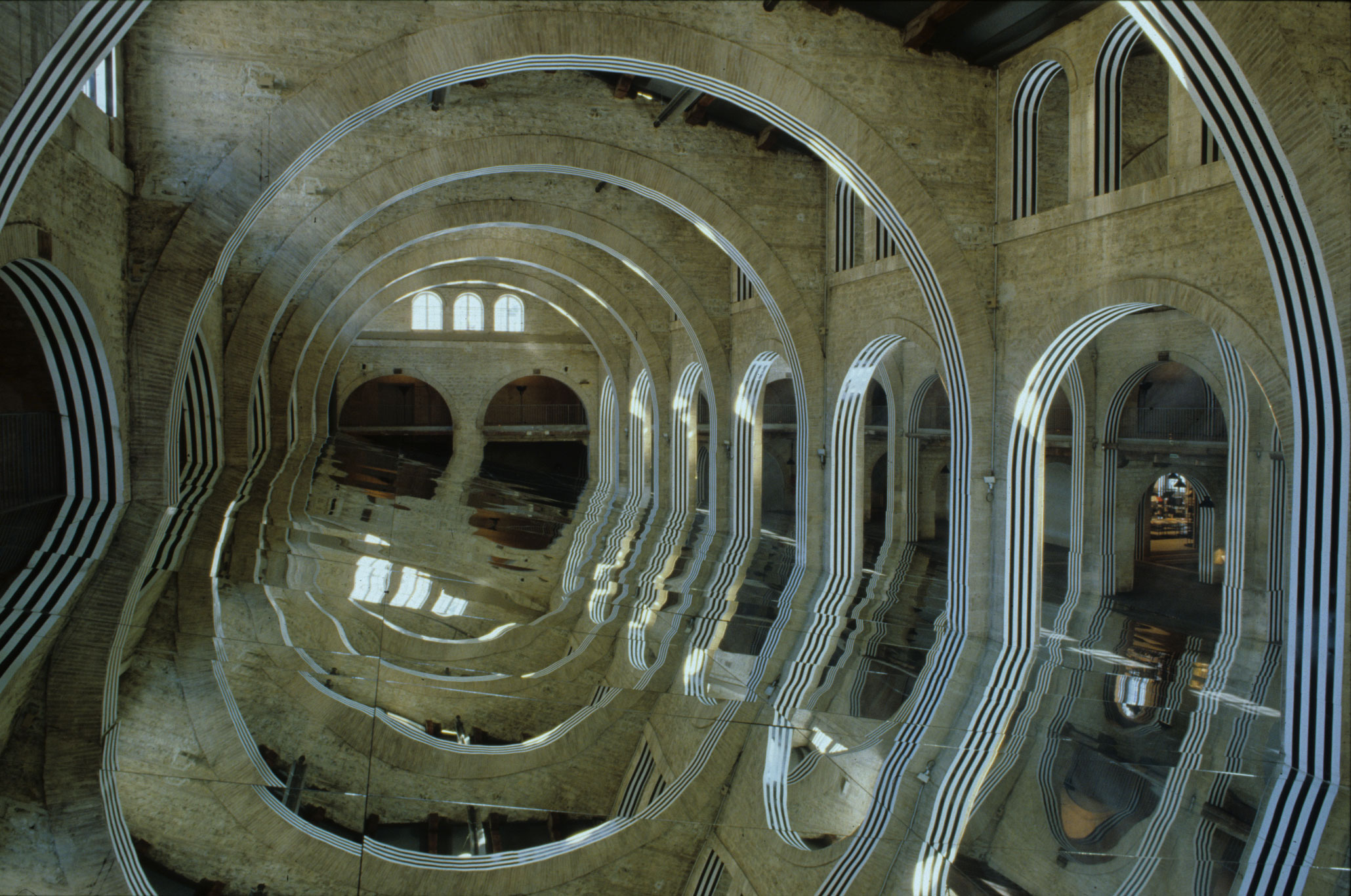 Un immense miroir oblique est installé dans la nef du Capc. Les arches en pierre du bâtiment sont recouvertes de bandes noires et blanches qui se reflètent sur le miroir