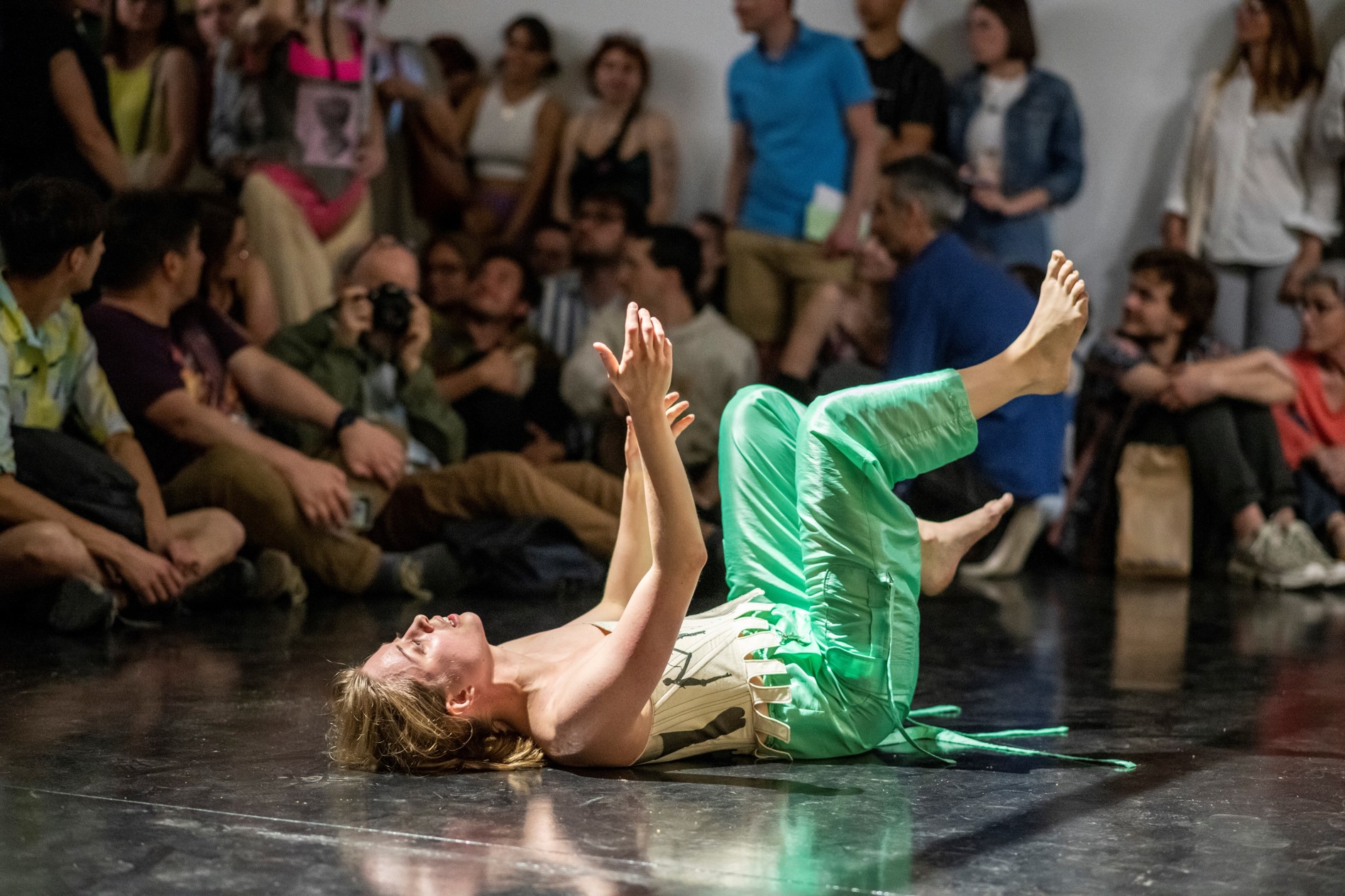 Une femme allongée sur le dos vêtu d'un pantalon vert et soyeux, les bras et les jambes vers le haut. Autour une assemblée regarde sa performance. 