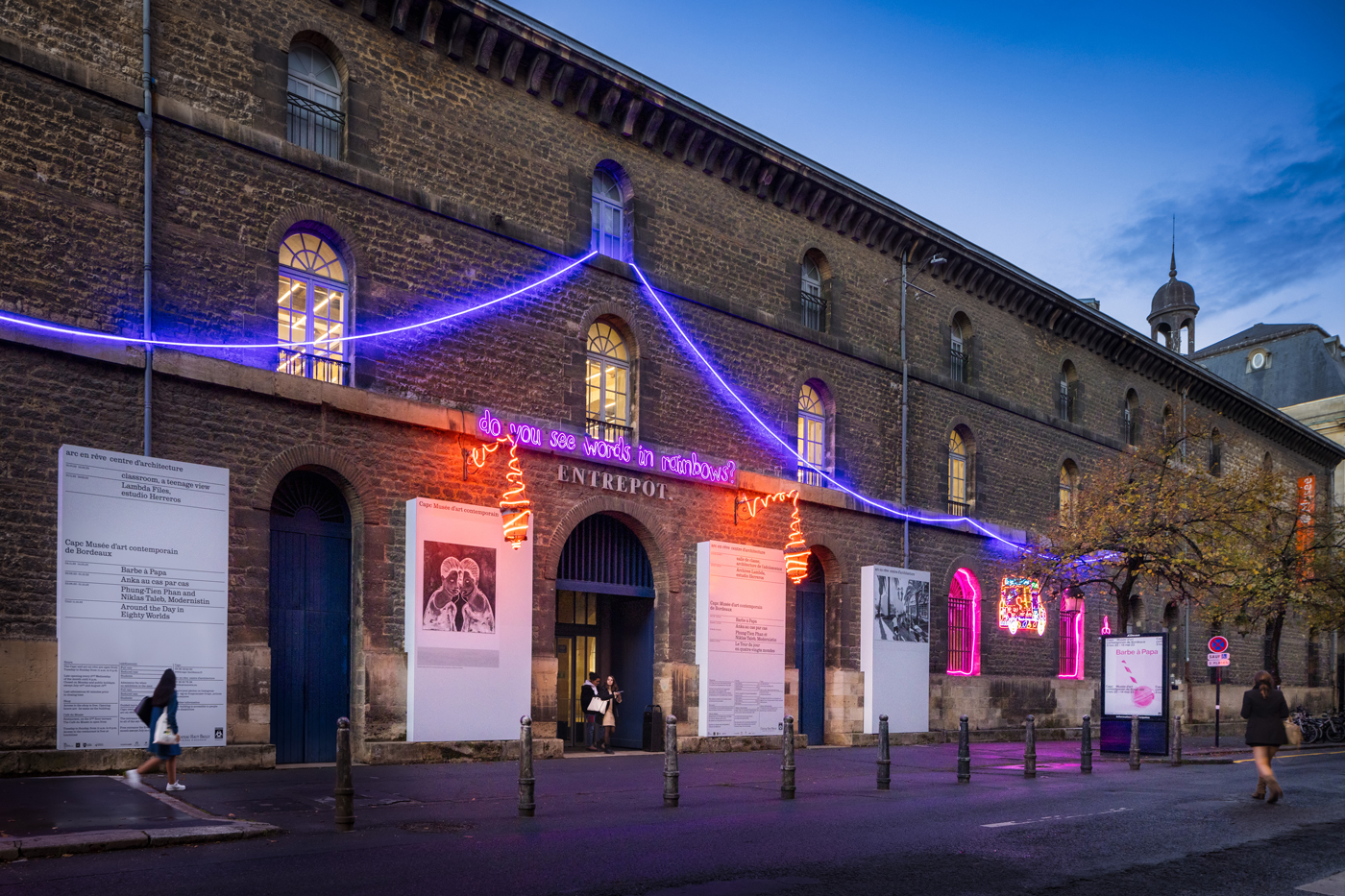 Sur la façade d'un bâtiment en pierre, de chaque côté de la grande porte centrale, des panneaux blancs portent des informations en lettrage noir. Certains panneaux sont agrémentés de visuel en couleur. Au-dessus un ensemble de néons colorés, comme des guirlandes, dont certains forme la phrase : Do you see words in...