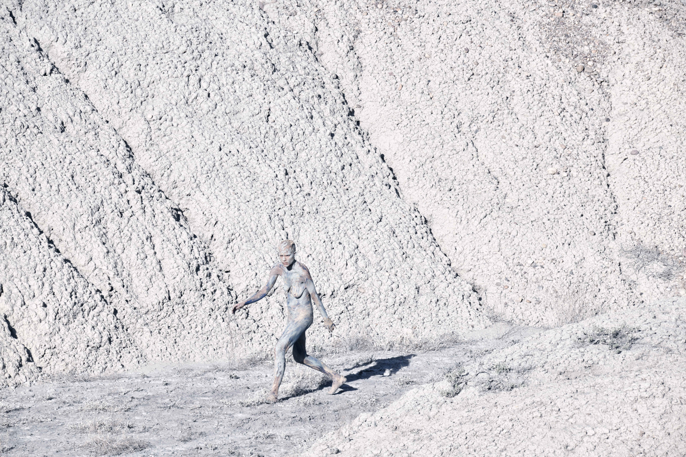 Une femme marche nue, au pied d'une montage. Le sol, la montagne et son corps sont recouvert d'une matière poudreuse et blanchâtre comme de la craie.