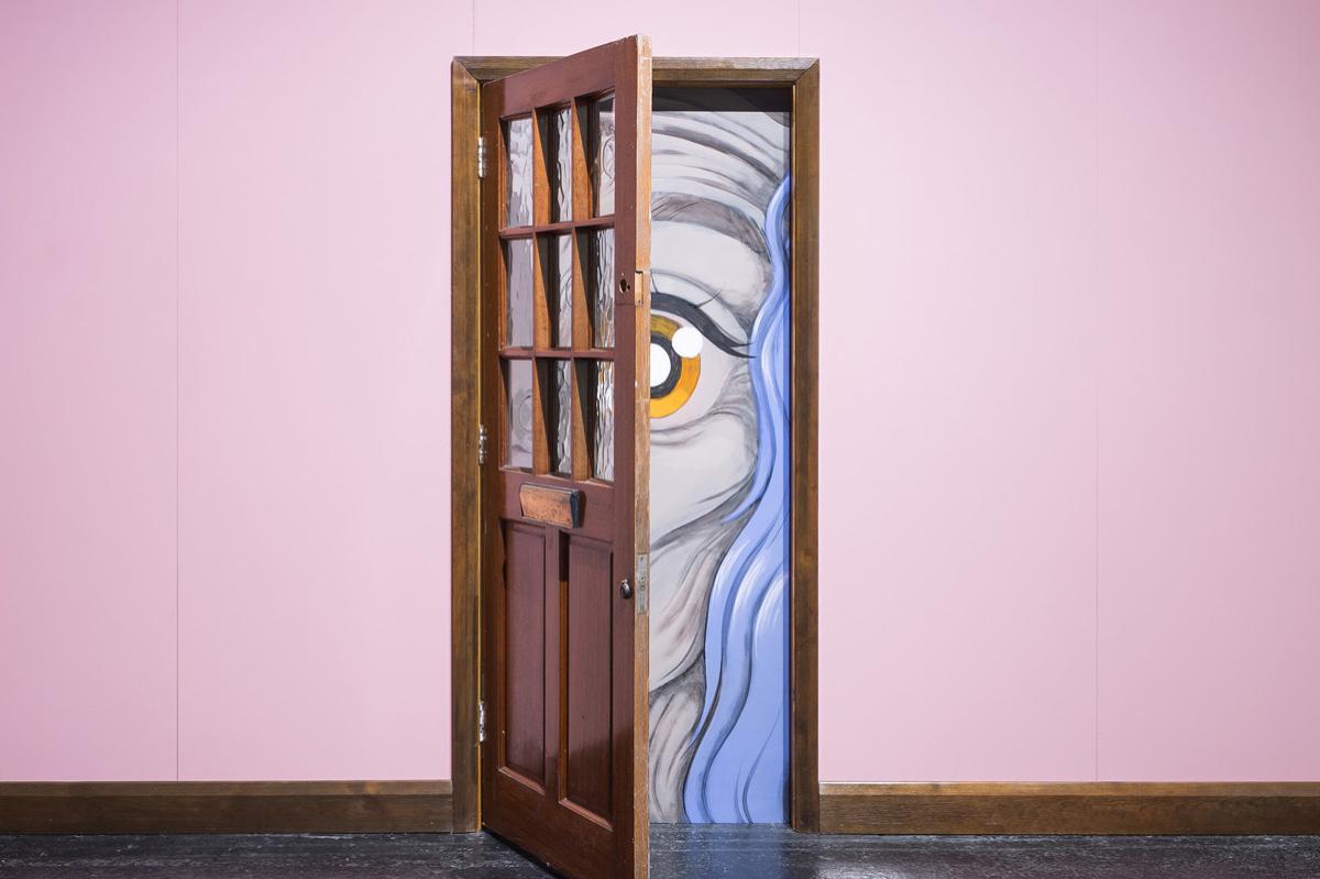 Au centre d'une salle d'exposition, une cloison peinte en rose avec au milieu une porte entre ouverte à travers laquelle on aperçoit en gros plan le visage émacié et ridé d'une femme aux cheveux bleus