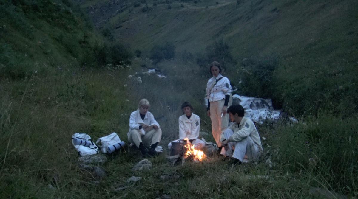 Quatre personnes vêtues de blanc sont autour d'un feu de camp dans la montagne. La nuit tombe.